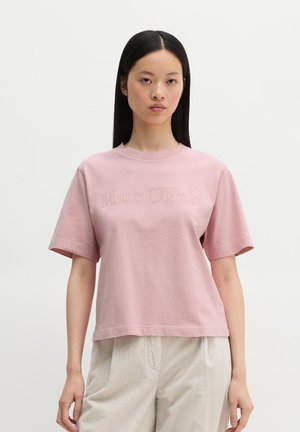 Kobieta z długimi ciemnymi włosami, ubrana w gładki jasnoróżowy t-shirt z wytłaczanym napisem oraz jasnobeżowe spodnie, stojąca na neutralnym tle.