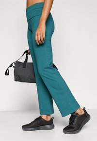 Τυρκουάζ παντελόνι γιόγκα με φαρδιά μέση, σε συνδυασμό με μαύρα αθλητικά παπούτσια με διαφανές ύφασμα και ένα σκούρο γκρι σακίδιο. Το ύφασμα φαίνεται λείο και ελαστικό.