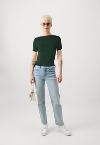 Mørkegrønn tettsittende t-skjorte, lyseblå rett-fot jeans, hvite joggesko, holder en liten kremfarget håndveske. Korte ermer, minimalistisk design.
