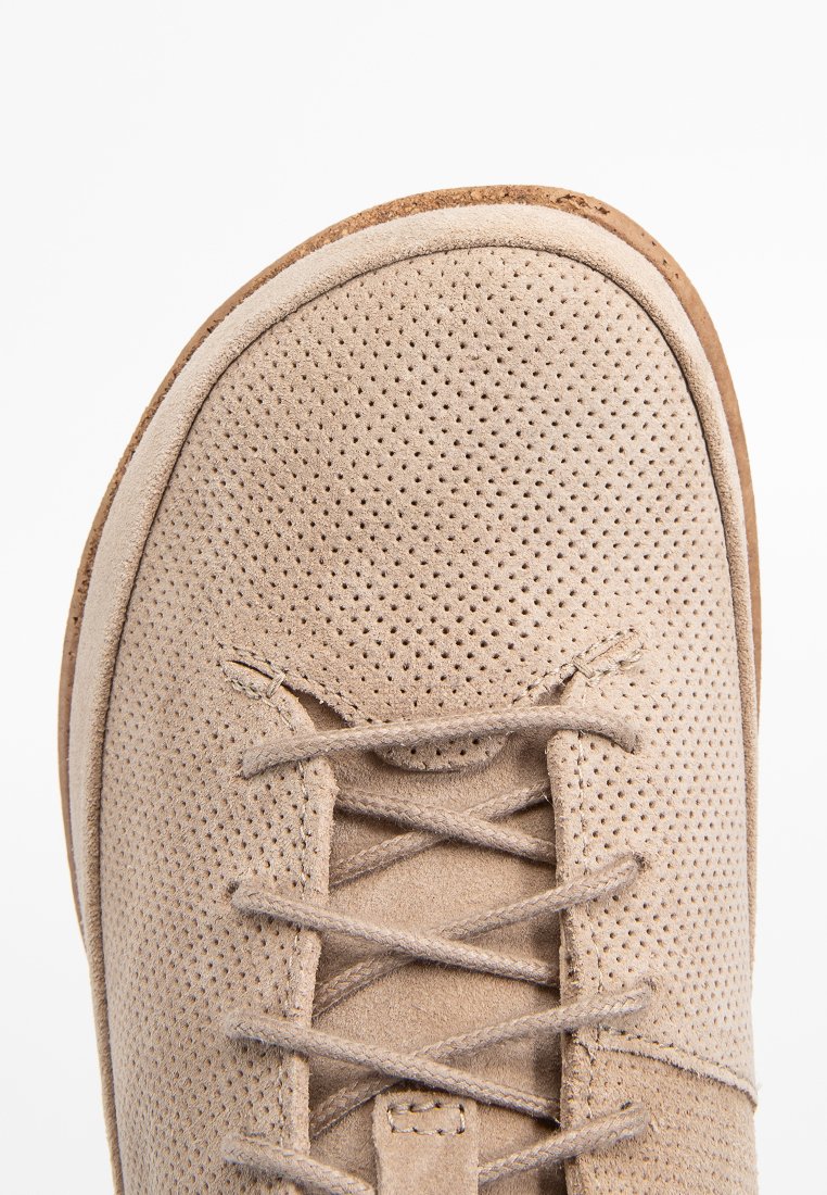 Close-up van de voorkant van een beige suède schoen met een geperforeerde textuur en beige veters, gedetailleerde stiksels zichtbaar op de zool en het bovenste deel.