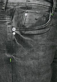 Jeans de mezclilla negra con herrajes plateados, diseño de cinco bolsillos y costura verde de contraste en la costura lateral. Acabado texturizado y desgastado.