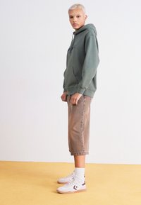 Grøn hættetrøje med frontlomme, parret med beige knælange shorts og hvide sneakers. Modellen står på et gult gulv mod en lys baggrund.