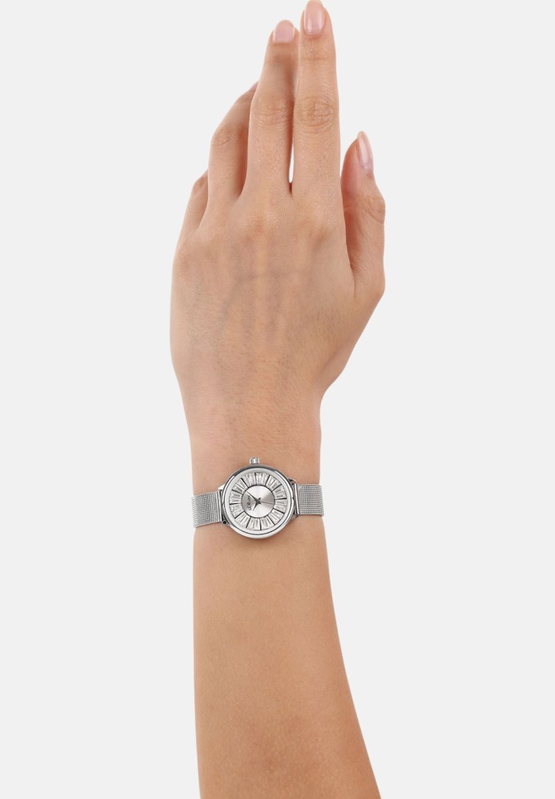 Silbernes Armband mit einem runden Zifferblatt, strukturiertem Mesh-Armband und Zifferblatt mit Sonnenschliff und Stundenmarkierungen. Die Hand, die die Uhr trägt, ist gehoben.