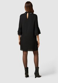 Schwarzes Kleid mit einem Schlüssellochausschnitt am Rücken, dreiviertellangen ausgestellten Ärmeln und transparenter Verzierung am Saum, kombiniert mit glänzenden schwarzen Knöchelstiefeln.