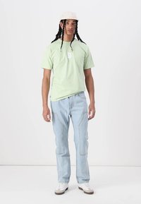 Világoszöld póló elülső grafikával, világoskék egyenes szabású farmer, bézs sapka és fehér sneaker barna részletekkel.
