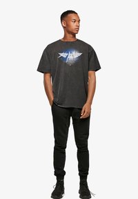 Grafisk svart bomulls t-skjorte med et blått og hvitt design i midten; kombinert med svarte joggebukser og sorte joggesko.