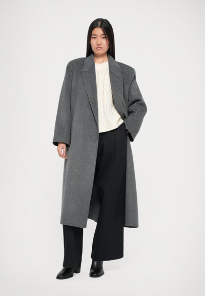 Mantou gri oversized cu guler tăiat, textură moale, mâneci largi, asortat cu un pulover tricotat crem și pantaloni negri cu croială largă.