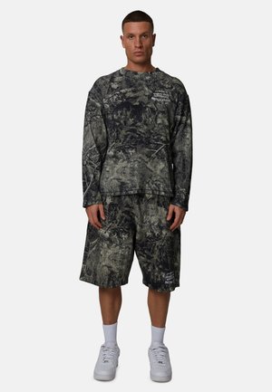 Uomo che indossa una maglia a maniche lunghe e pantaloncini lunghi mimetici neri e grigi abbinati, calzini bianchi e scarpe da ginnastica bianche, in piedi contro uno sfondo semplice.