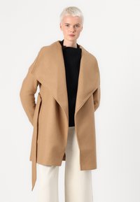 Manteau en laine beige avec un col châle surdimensionné, ouvert devant, ceinture à nouer et poches avant. Porté par-dessus un pull en maille noir.