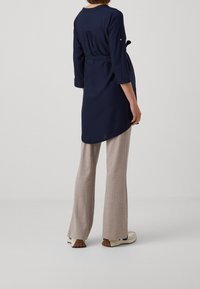 Navy long tunic with a cinched waist, rounded hem, and three-quarter sleeves paired with beige wide-leg pants and cream shoes.