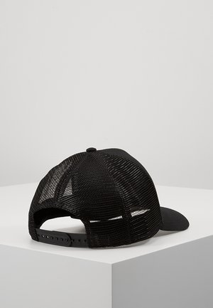 Czarna siateczkowa czapka typu trucker z regulowanym zapięciem na zatrzask, leżąca na białej powierzchni na tle jednolitego jasnoszarego tła.