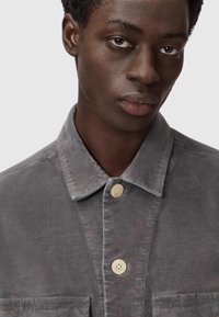 Grijze ribfluwelen jas met een puntige kraag, twee voorkktten en beige knopen. De stof heeft een zachte, gestructureerde uitstraling.