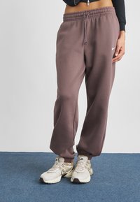 Violetiniai sportiniai šortai su elastinga juosme ir rankovėmis, su baltu Nike logotipu. Derinami su bežiais ir rudais sportbačiais ant mėlynos dangos.