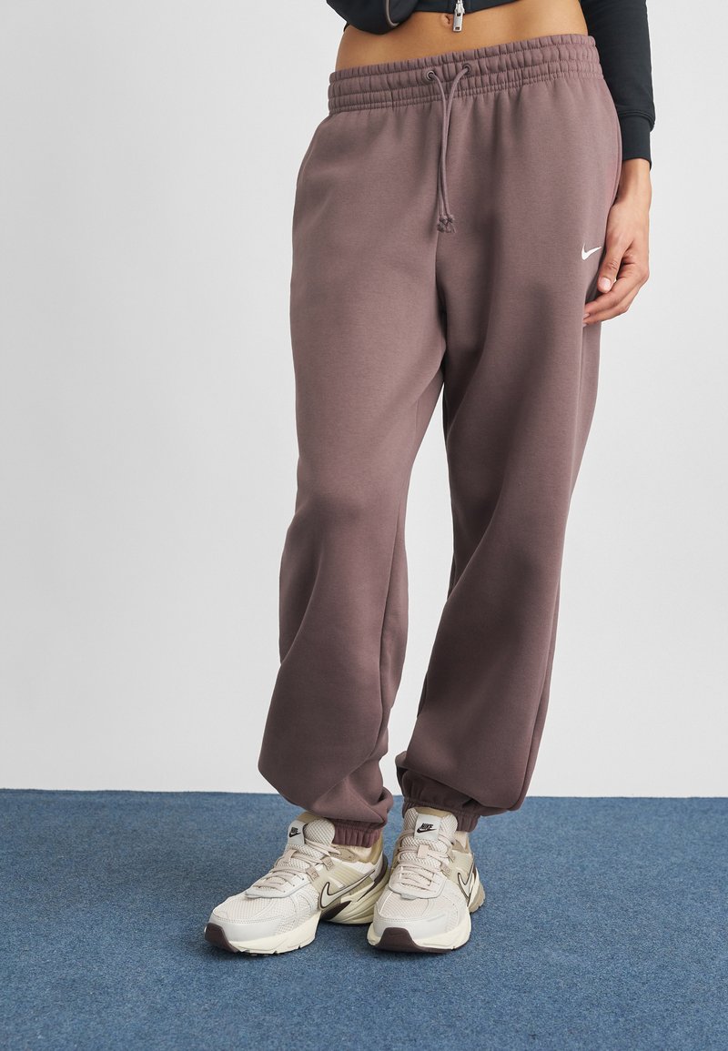 Violetiniai sportiniai šortai su elastinga juosme ir rankovėmis, su baltu Nike logotipu. Derinami su bežiais ir rudais sportbačiais ant mėlynos dangos.