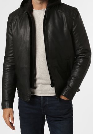 Mężczyzna ubrany w czarną skórzaną kurtkę na jasnoszarej koszuli i ciemnoniebieskich dżinsach, z jedną ręką w kieszeni i częściowo rozpiętym zamkiem.