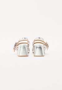 Sandali metallici argentati con tacco a blocco, fascette alla caviglia e dettagli decorativi a forma di fiocco. Texture liscia con una leggera lucentezza.