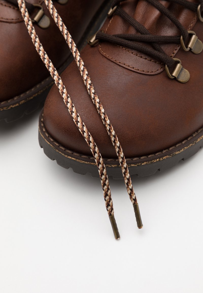 Braune Lederwanderstiefel mit strukturierter Oberfläche, ausgestattet mit braunen und beigefarbenen geflochtenen Schnürsenkeln und Metallösen. Schwarze Gummisohle mit Profil.