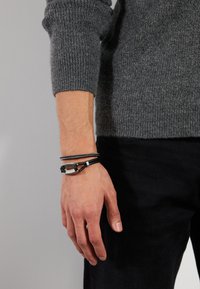 Sort læderarmbånd med metal detaljer, struktureret grå striktrøje og mørkeblå jeans. Hånd positioneret ved siden, der viser tilbehøret.