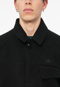 Cappotto nero in misto lana con colletto strutturato, chiusura a bottoni e tasca frontale. Presenta un logo discreto sul lato sinistro.