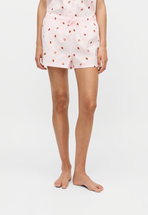 Frau trägt hellrosa Shorts mit kleinen roten Erdbeer-Prints und einem passenden Shirt, steht barfuß vor einem schlichten Hintergrund.