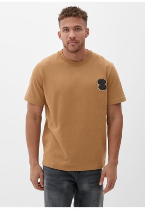 Bruine katoenen T-shirt met korte mouwen, ronde hals en een zwart geborduurd logo op de linkerkant van de borst. Model draagt een donkere spijkerbroek.