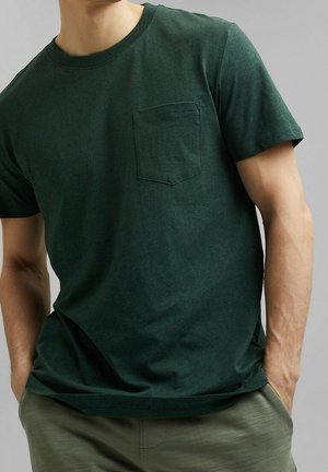 Mann trägt dunkelgrünes Kurzarm-T-Shirt mit Brusttasche, Hände in den Taschen und olivgrüne Hose vor grauem Hintergrund.