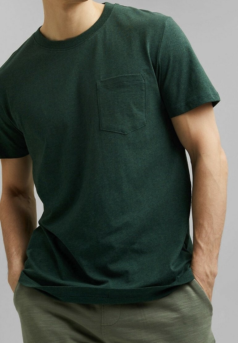 Homme portant un t-shirt manches courtes vert foncé avec poche poitrine, les mains dans les poches, et un pantalon vert olive sur fond gris.