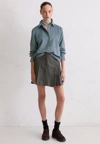 Helles blaues Hemd mit hochgekrempelten Ärmeln, grauer plissierter Rock, schwarze kniehohe Socken und braune Slipper vor einem neutralen Hintergrund.