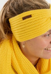 Fascia gialla lavorata a maglia con un design attorcigliato e un'etichetta in legno. Accompagnata da una sciarpa gialla coordinata e strutturata. Orecchino a forma di foglia visibile.