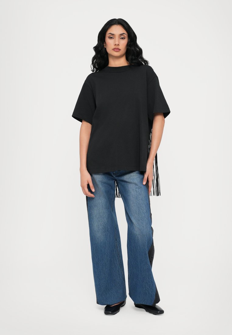 T-shirt noir oversize à manches courtes, avec des détails frangés sur le côté. Associé à un jean en denim bleu à jambes larges.