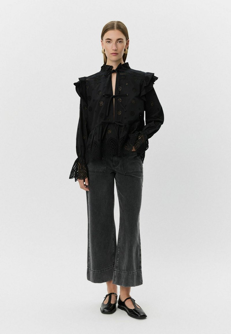 Blusa negra con mangas con volantes y detalles recortados, combinada con culottes de mezclilla negra de talle alto y zapatos planos negros.