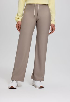 Person trägt beige weit geschnittene Hosen mit Kordelzug am Bund, weiße Sneakers und ein hellgelbes Langarmshirt vor einem einfarbigen Hintergrund.