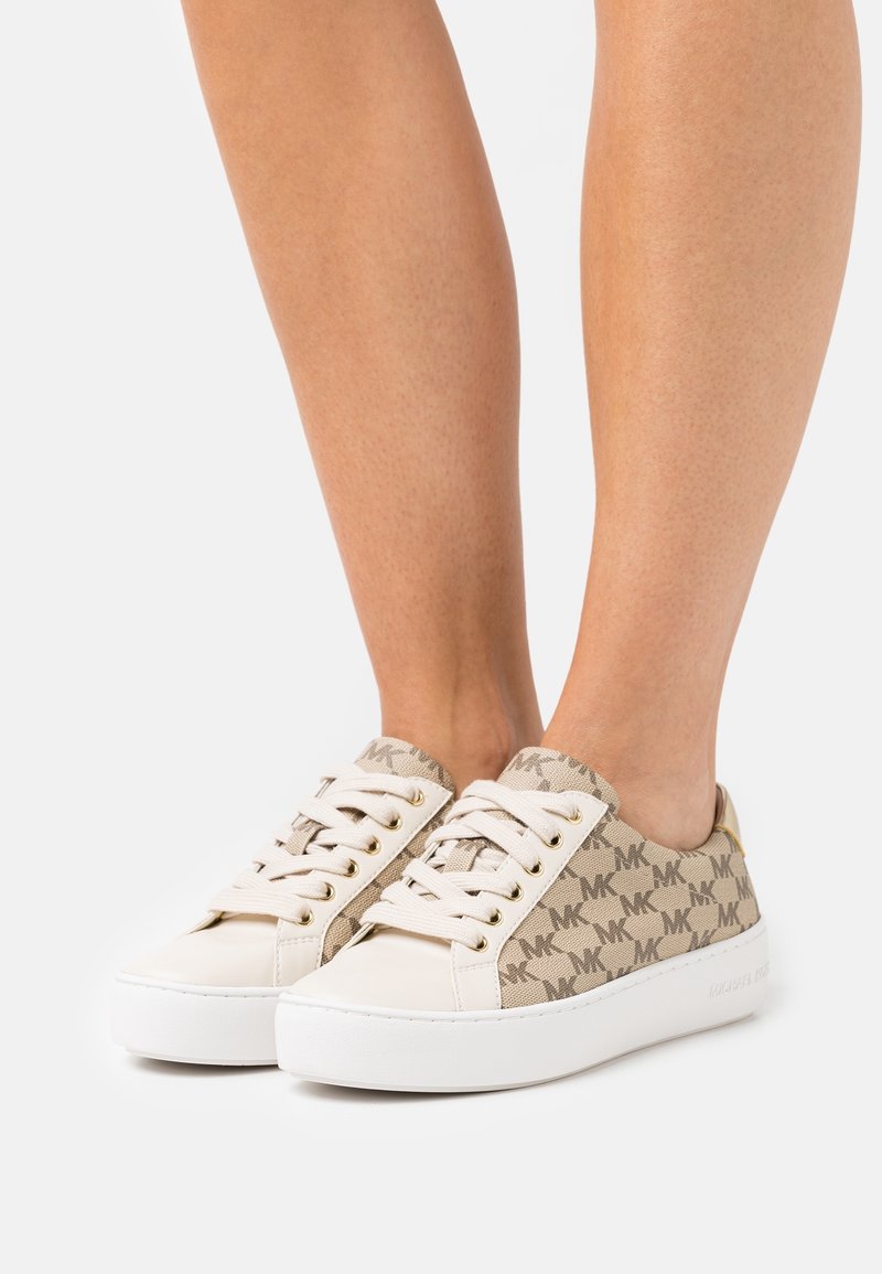 Jambes portant des baskets beige Michael Kors avec des lacets blancs, des semelles blanches et des panneaux en tissu avec le monogramme MK.