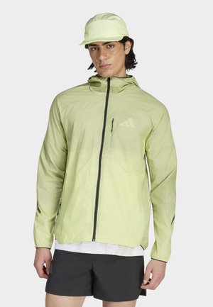 Mann trägt hellgrüne Kapuzenjacke mit Frontreißverschluss, passende Mütze, schwarze Shorts und weißes Hemd vor einfarbigem Hintergrund.