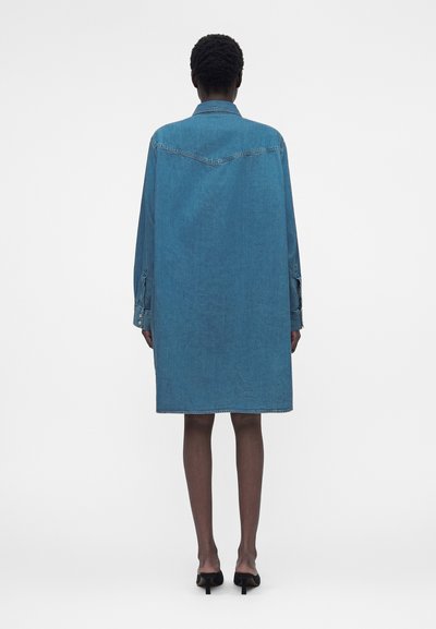 Άτομο που στέκεται με την πλάτη, φορώντας ένα φαρδύ μπλε τζιν φόρεμα που φτάνει μέχρι το γόνατο και μαύρα kitten heels, σε απλό λευκό φόντο.