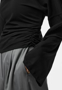Top nero a maniche lunghe con dettaglio arricciato sui lati, abbinato a pantaloni grigi a righe sottili a vita alta, che mostrano texture morbide e colori a contrasto.