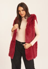 Burgundy faux fur vest with a hood, featuring a full length and front opening. Worn over a fitted beige top and black pants.
