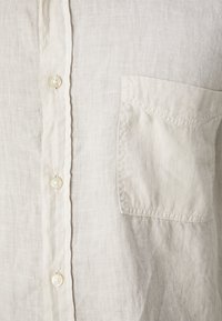 Camisa de lino gris claro con parte delantera abotonada, que presenta botones perlados y un bolsillo de parche en el lado izquierdo. Tela texturizada.