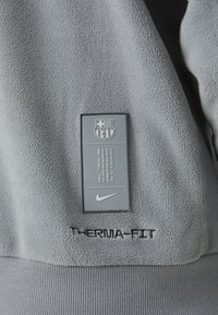 Γκρι φλις πουλόβερ με υφή στην επιφάνεια, διαθέτει ορθογώνια ετικέτα με το λογότυπο της FC Barcelona και τη λέξη "THERMA-FIT" κεντημένη σε μαύρο χρώμα.