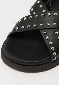 Svart slidesandal med kryssede stropper som har sølvnitter langs kantene. Teksturert materiale, flat såle med en glatt svart finish.