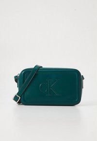 Žalsvai mėlyna odinė rankinė per petį stačiakampio formos, su įspaustu „cK“ logotipu, sidabrinės spalvos detalėmis ir reguliuojamu dirželiu. Lygios faktūros.