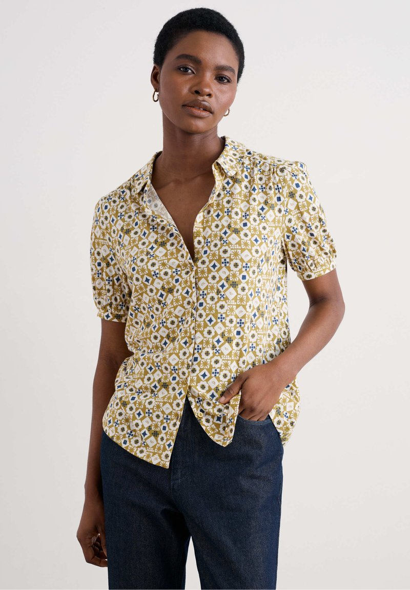 Camicia a manica corta con bottoni di colore giallo, decorata con motivi floreali geometrici blu, in materiale di cotone e con un taglio relaxed. Dettagli includono un colletto a punta.