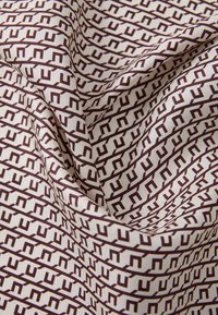 Textil, amelynek krémszínű háttere van, barna geometrikus mintával, amely stilizált betűkre emlékeztet, textúrált megjelenést kölcsönözve.