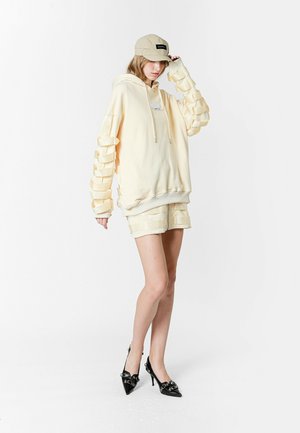 Sudadera oversized amarilla clara con mangas texturizadas, combinada con pantalones cortos a juego. La modelo lleva una gorra beige y zapatos de tacón negros.