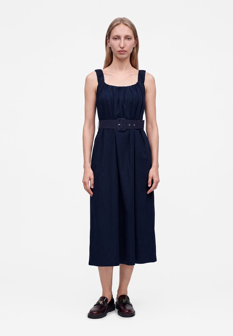 Femme debout portant une robe midi sans manches bleu marine avec un col carré, une taille ceinturée et des mocassins foncés sur un fond clair uni.