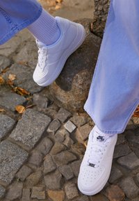 Person som bär vita Nike Air Force 1-sneakers med vita strumpor och blå byxor står på kullerstensbeläggning nära en sten.
