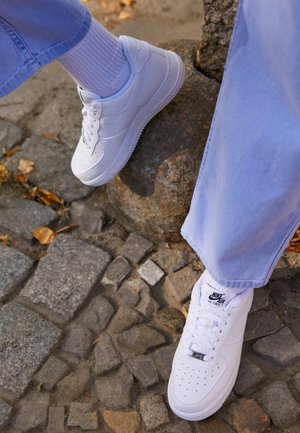 Personne portant des baskets blanches Nike Air Force 1 avec des chaussettes blanches et un pantalon bleu, debout sur un pavé en pierre près d'un rocher.