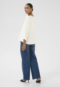 Top blanc ample avec des manches larges, associé à un jean bleu à jambes larges. Le mannequin se tient de côté, mettant en valeur un style décontracté. Chaussures à talons noires.