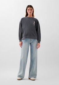 Felpa grigia oversize con collo rotondo e dettaglio del logo, abbinata a jeans larghi azzurri e sneakers beige.