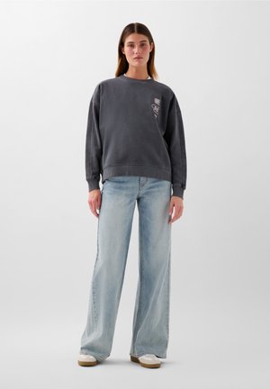 Felpa grigia oversize con collo rotondo e dettaglio del logo, abbinata a jeans larghi azzurri e sneakers beige.